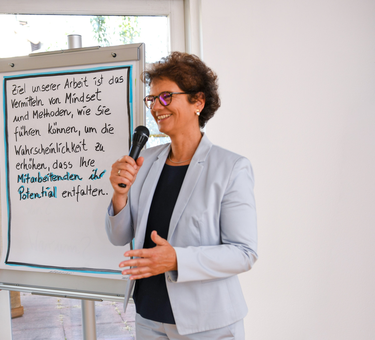 PermaTeam – Judith Eggersdorfer steht vor einem Flipchart mit einem Mikrophon in der Hand. Sie lächelt und gestikuliert beschreibend. Auf dem Flipchart steht handschriftlich: Ziel unserer Arbeit ist das Vermitteln von Mindset und Methoden, wie Sie führen können, um die Wahrscheinlichkeit zu erhöhen, dass Ihre Mitarbeitenden ihr Potentiall entfalten.